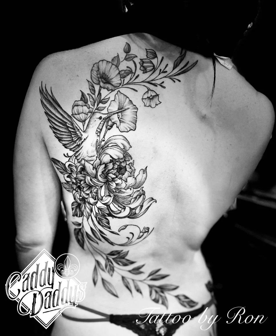 Black and white photo of a person's bare back with a large floral tattoo featuring a hummingbird, flowers, and leaves, with text reading 'Caddy Daddys' and 'Tattoo by Ron'.