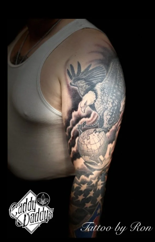 A person with a sleeve tattoo on their arm featuring an eagle, a globe, and American flag elements.