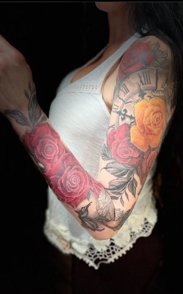 Tattooed woman's arm with colorful floral and clock designs, wearing a white sleeveless top.