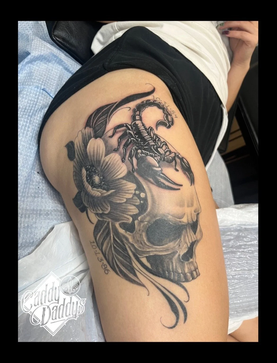 Tattoo of a skull with a scorpion on top and a flower near the skull's eye socket on a person's upper thigh.
