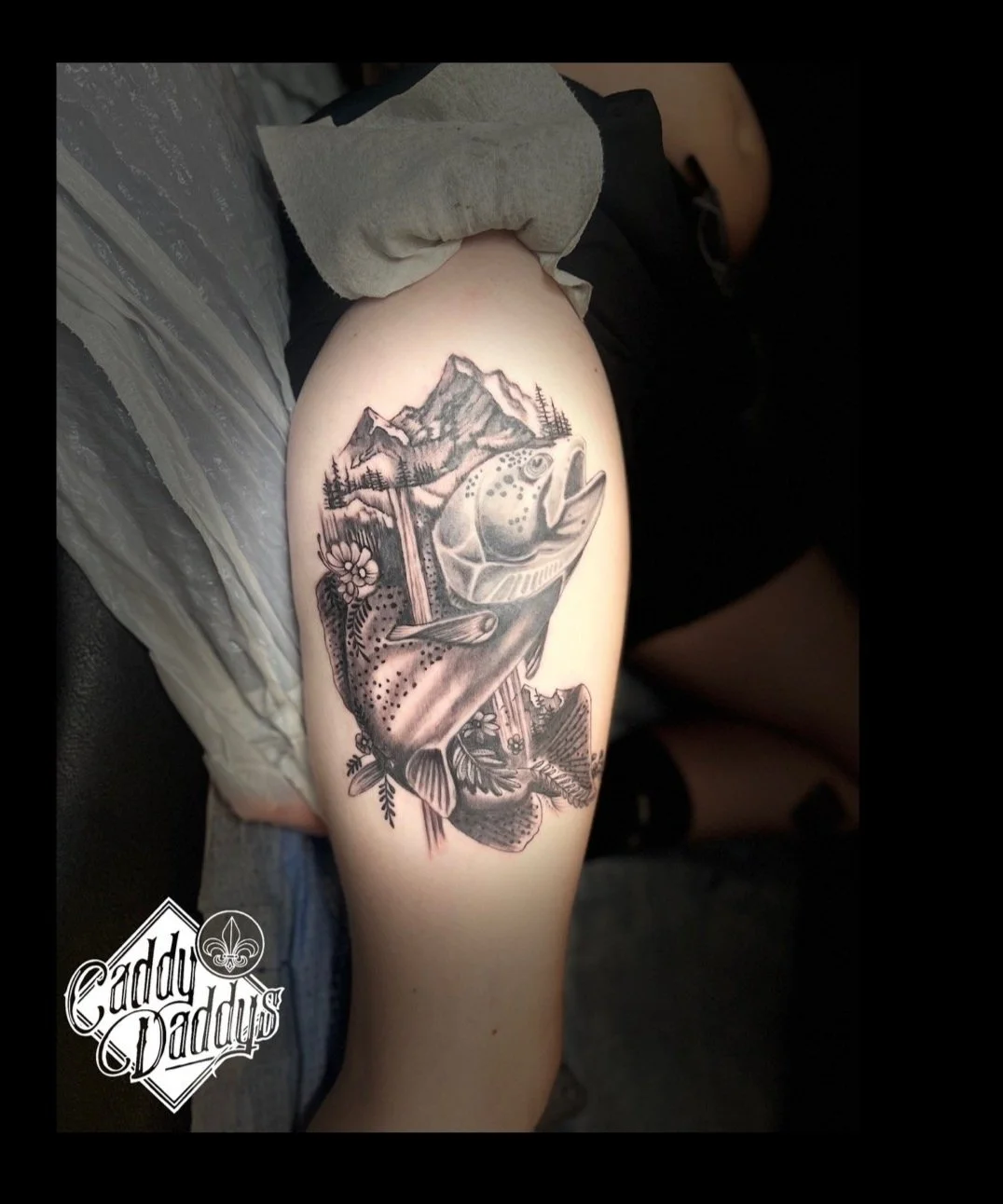 Tattoo of a mountain landscape with a fish, trees, and flowers on a person's upper thigh.
