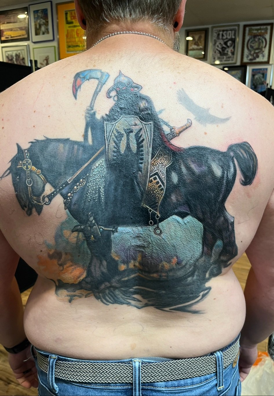 A detailed tattoo on a person's back depicting a black horse with armor and a knight riding it, holding a scythe. The background includes clouds and a bird flying.