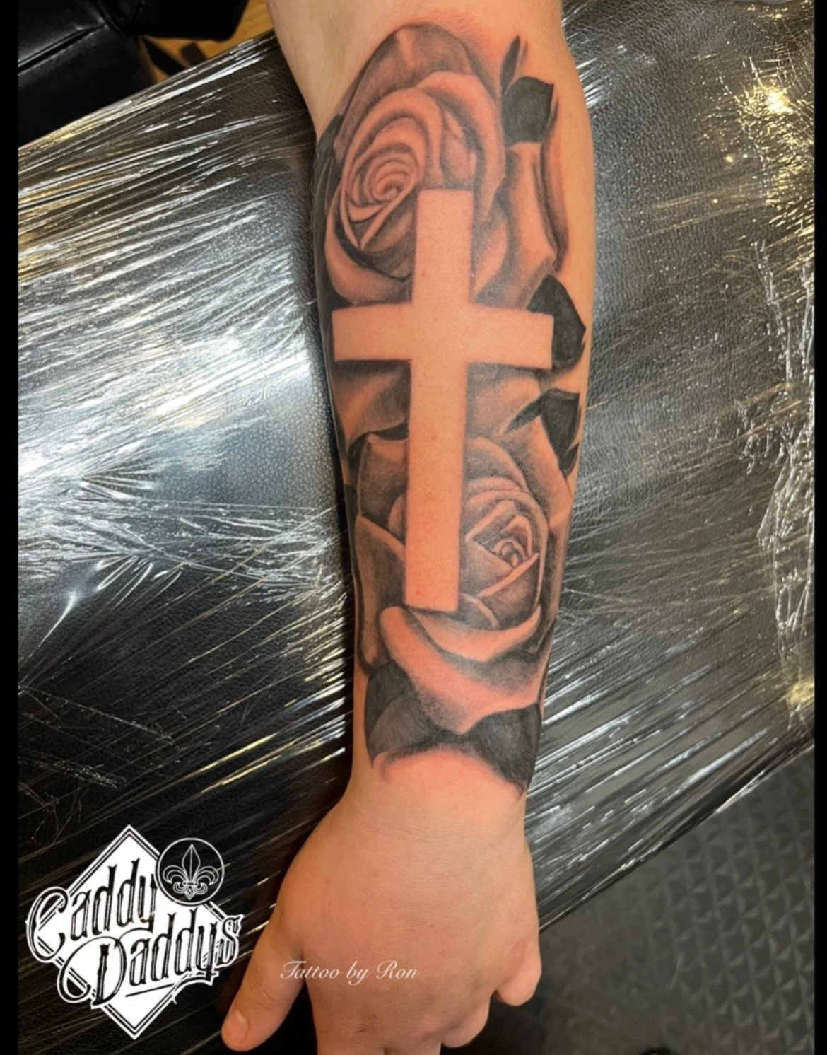 Tattoo of a large cross overlaid on detailed black and gray roses on an arm. The tattoo includes a smaller rose near the wrist and another near the elbow.