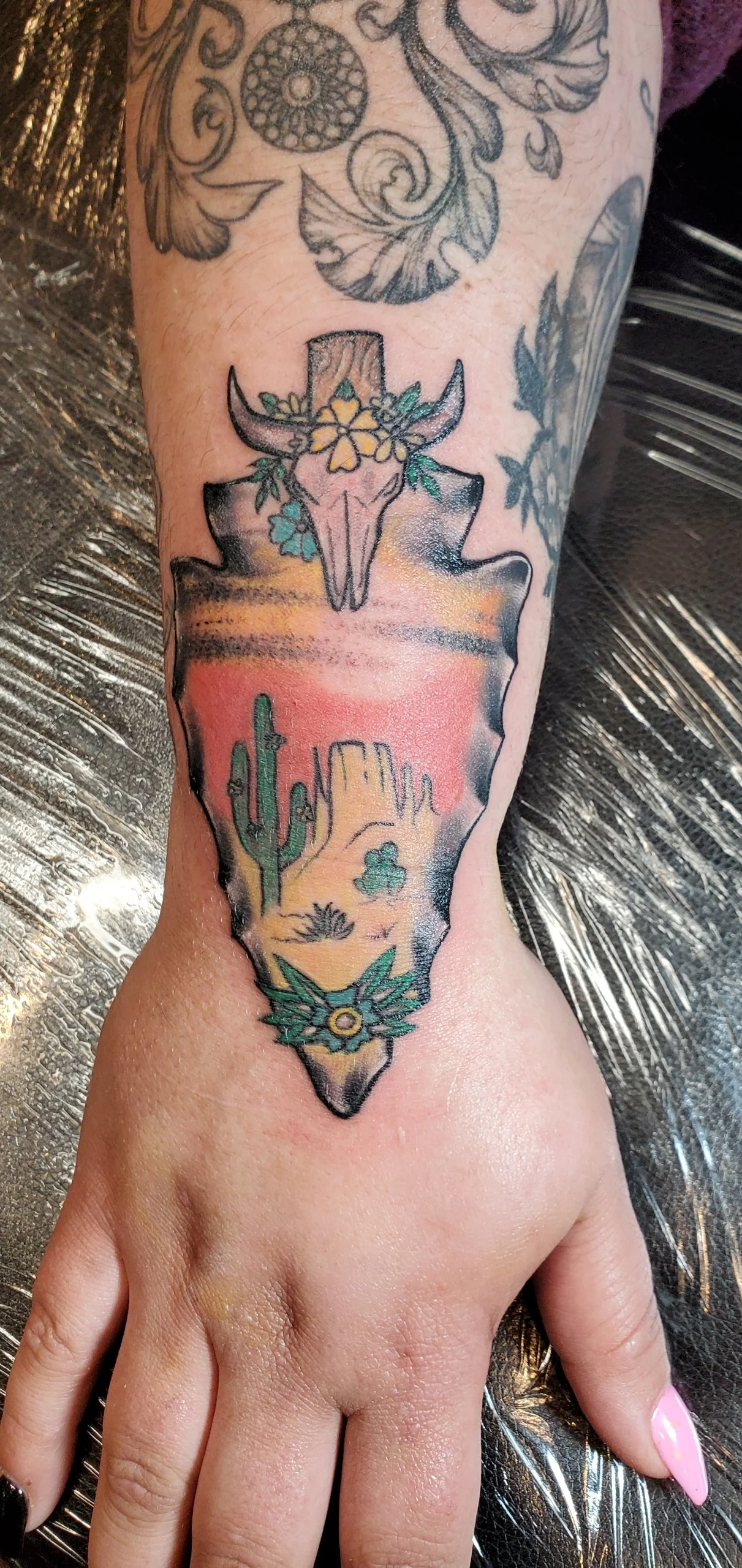 Tattoo of a desert scene with cacti and rock formations inside an outlined shape, a skull with deer antlers and floral decoration above, and a decorative element below, done by the best tattoo shop in Redding
