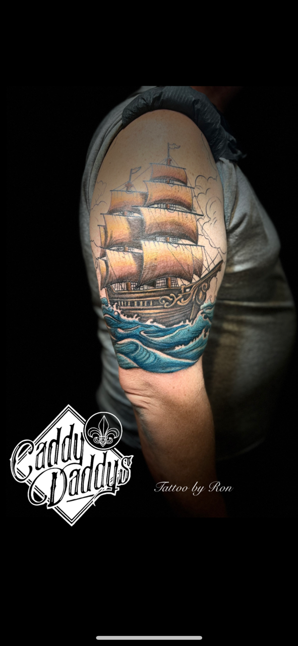 Tattoo of a sailing ship on a person's upper arm with waves below and clouds in the background.