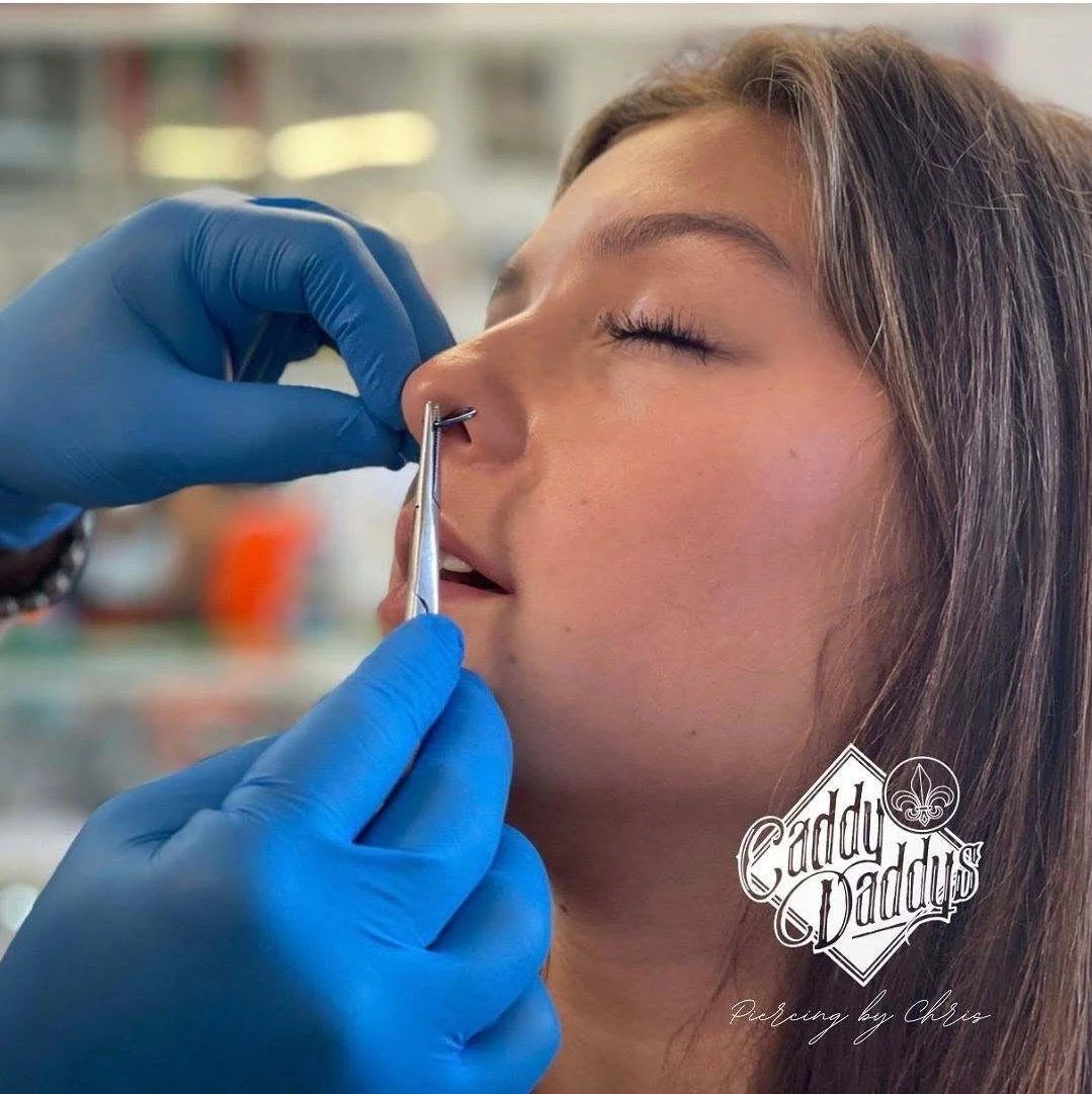 A woman receiving a nose piercing from a person wearing blue gloves in a clinical setting.