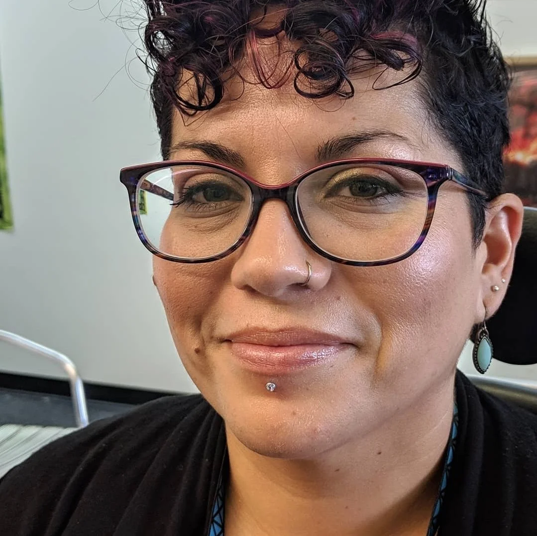 Close-up of a woman with short curly dark hair, wearing glasses, earrings, and a nose ring from local piercing shop Caddy Daddy's