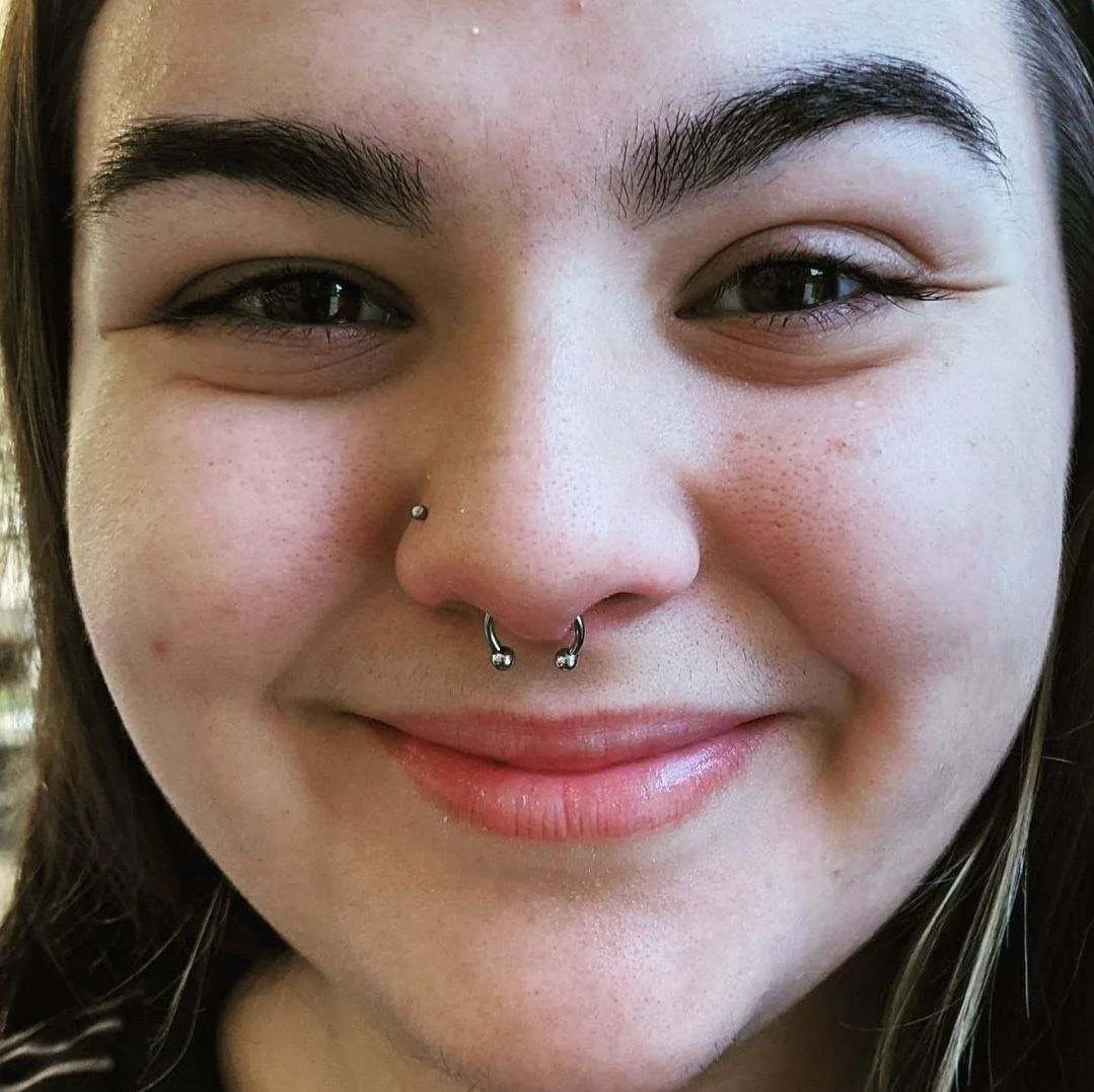 Close-up of a smiling woman with dark eyebrows and nose piercings, including a septum ring and a nostril stud.