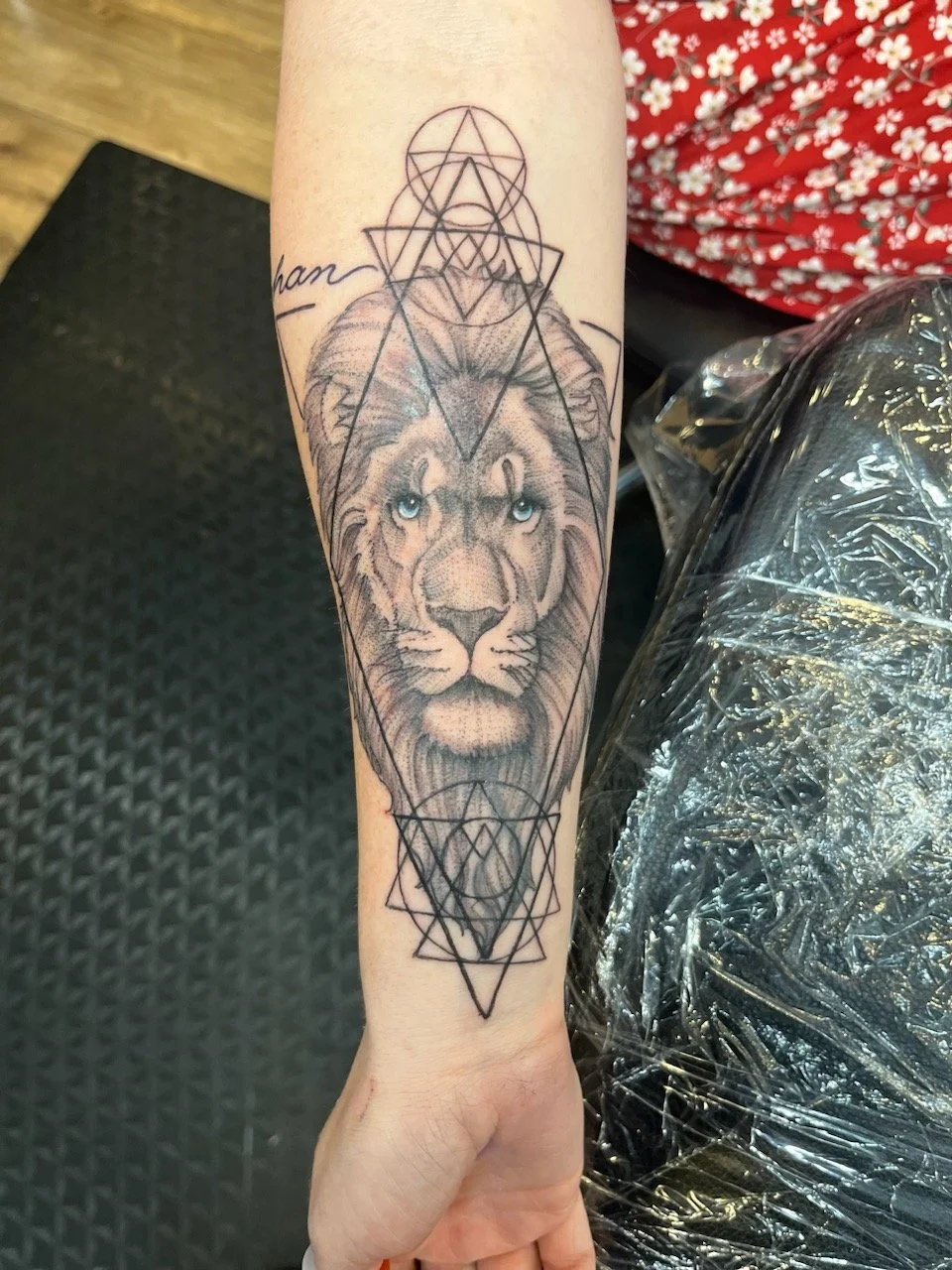 Tattoo of a lion's face with blue eyes, surrounded by geometric shapes on a person's forearm.