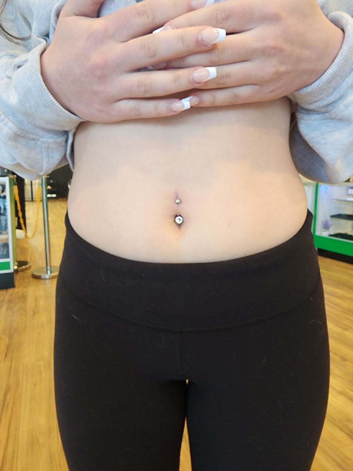 Close-up of a woman's midsection showing her belly with a navel piercing, wearing black pants and a gray sweatshirt, with her hands resting on her chest.