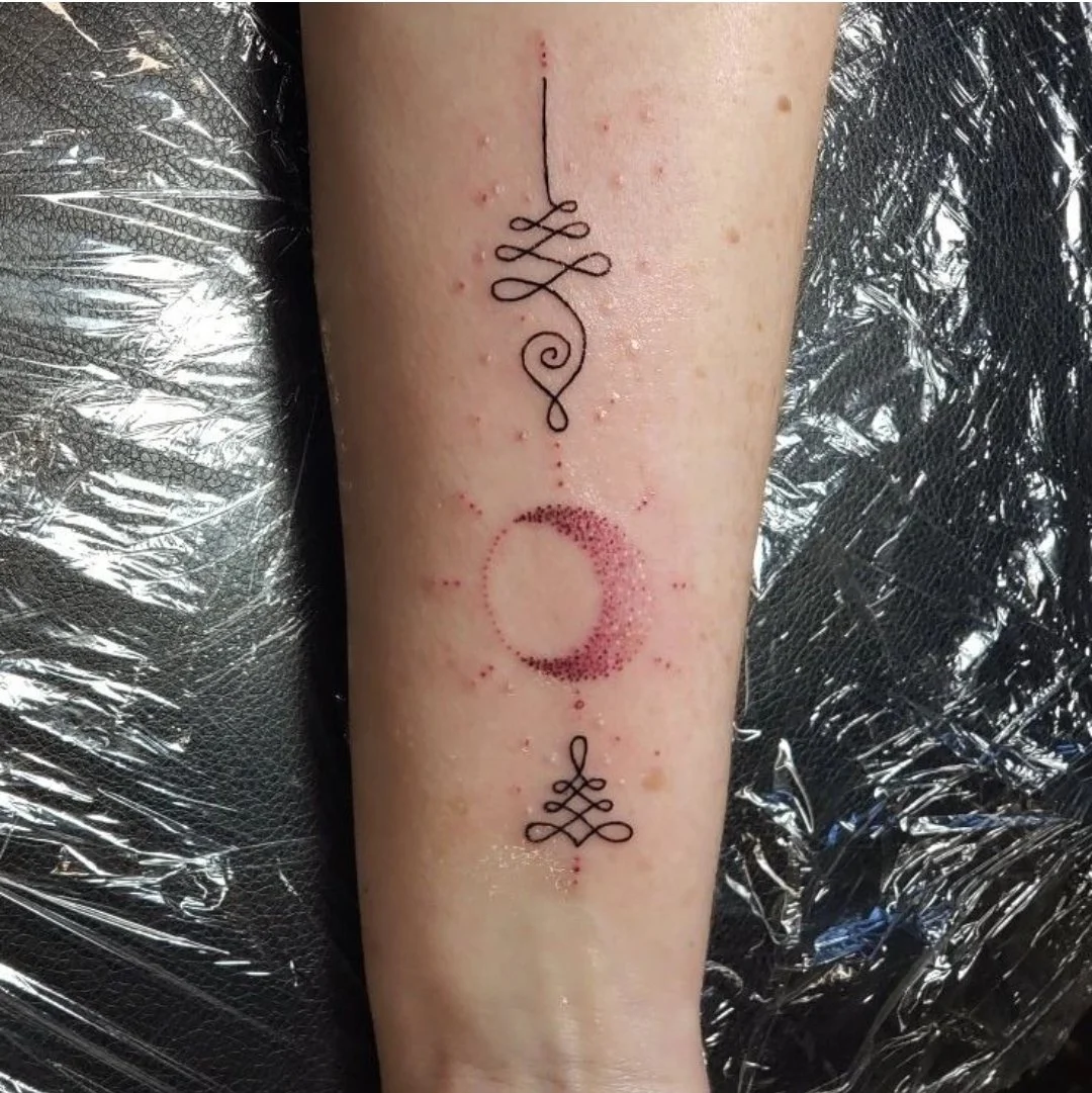 Tattoo on forearm, featuring a black ink design with a looped line at the top, a dotted pink circle in the middle, and a looped line at the bottom.