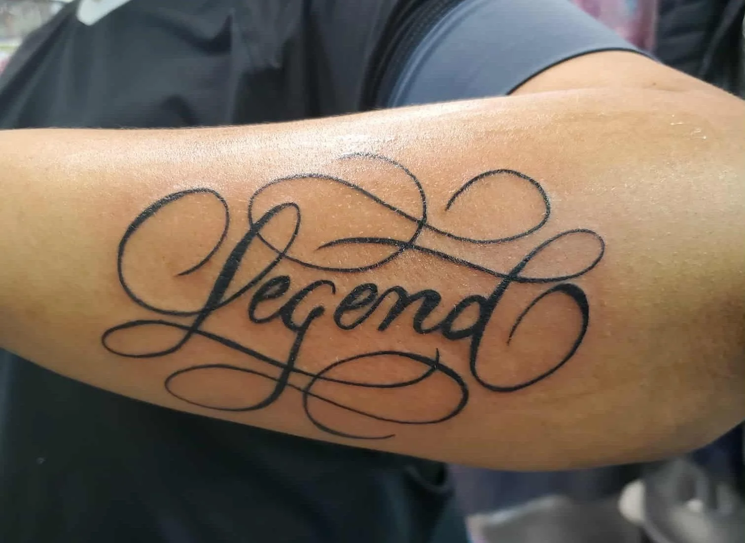 Close-up of a tattoo on a person's arm that reads 'Legend' in cursive script with decorative swirls.
