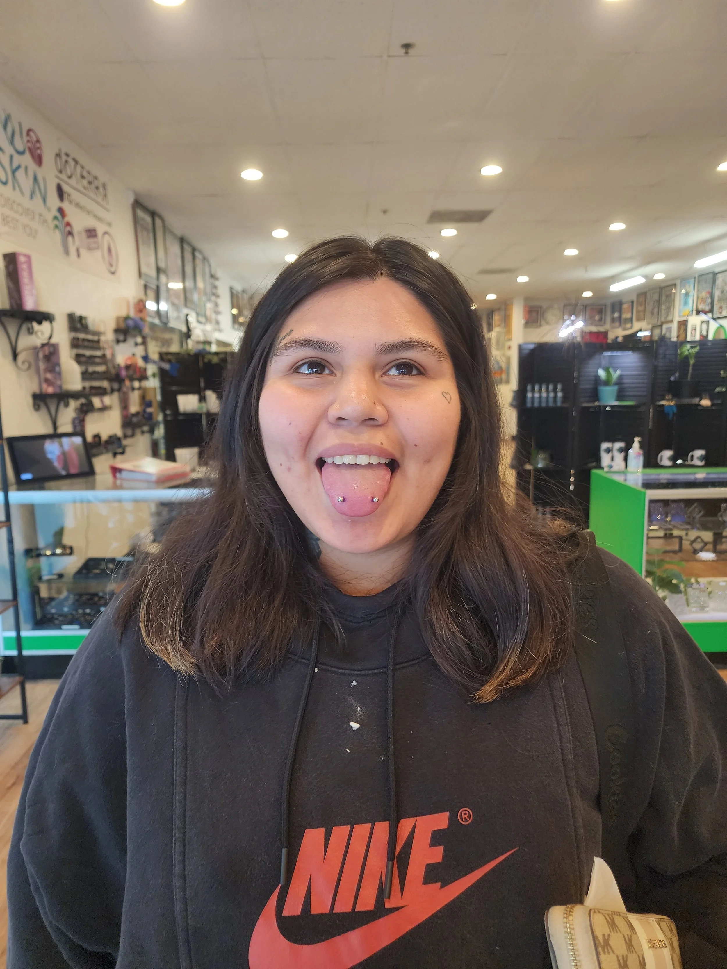 A young woman with dark hair, tongue pierced, wearing a black Nike hoodie, smiling in a retail store.