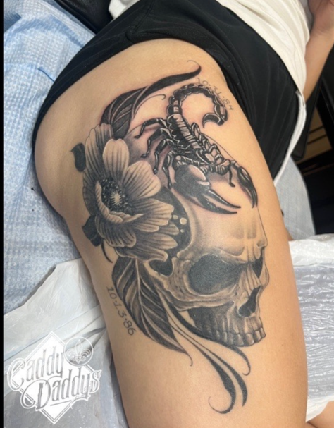 upper thigh tattoo of a skull with flowers and a scorpion