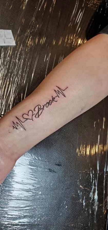 Tattoo on person's forearm with a heart symbol, the words 'My Brook' and a heartbeat line.