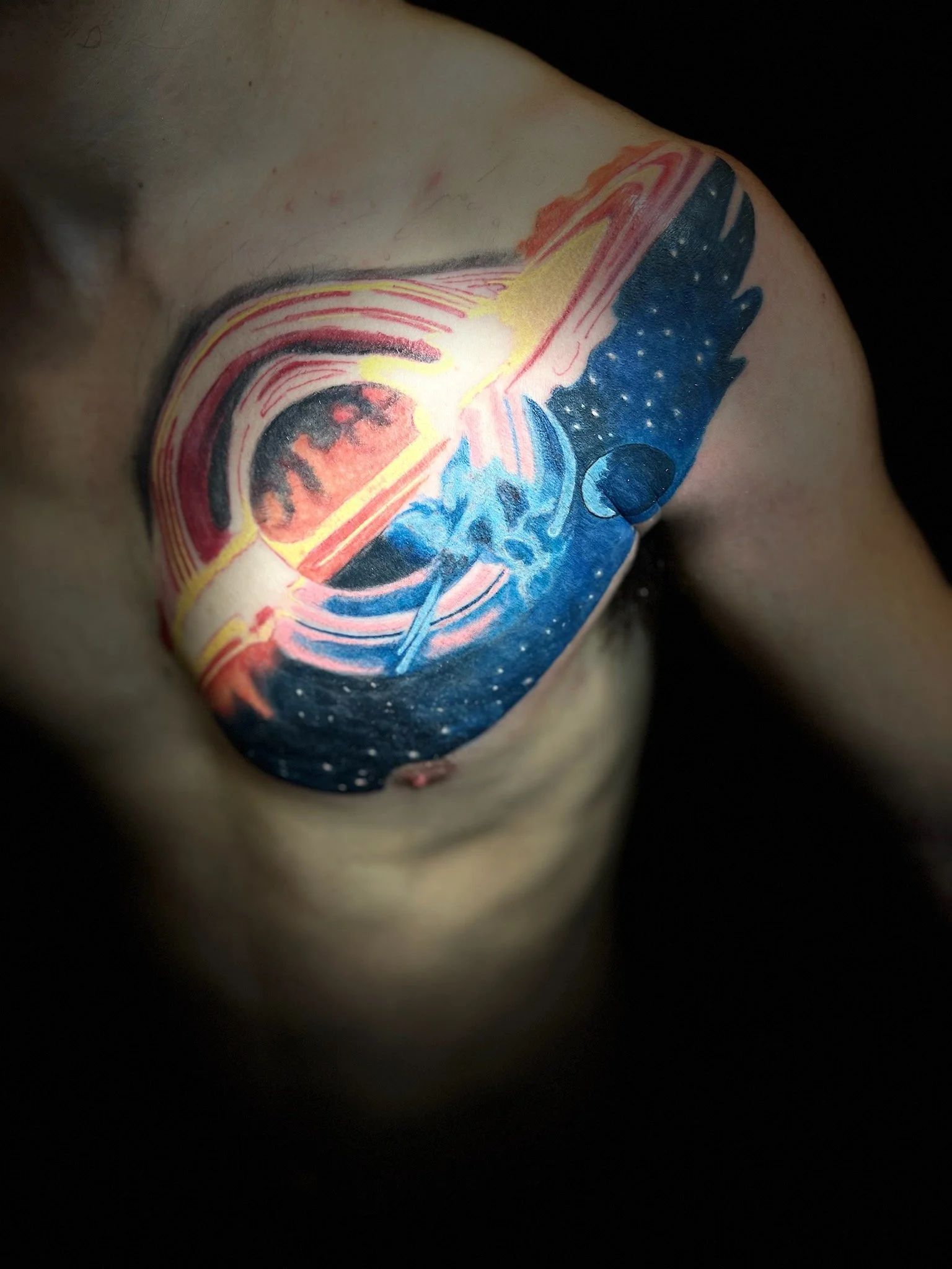 Colorful shoulder tattoo depicting an eye and cosmic elements, including stars and planets, with vibrant colors of red, blue, yellow, and black.