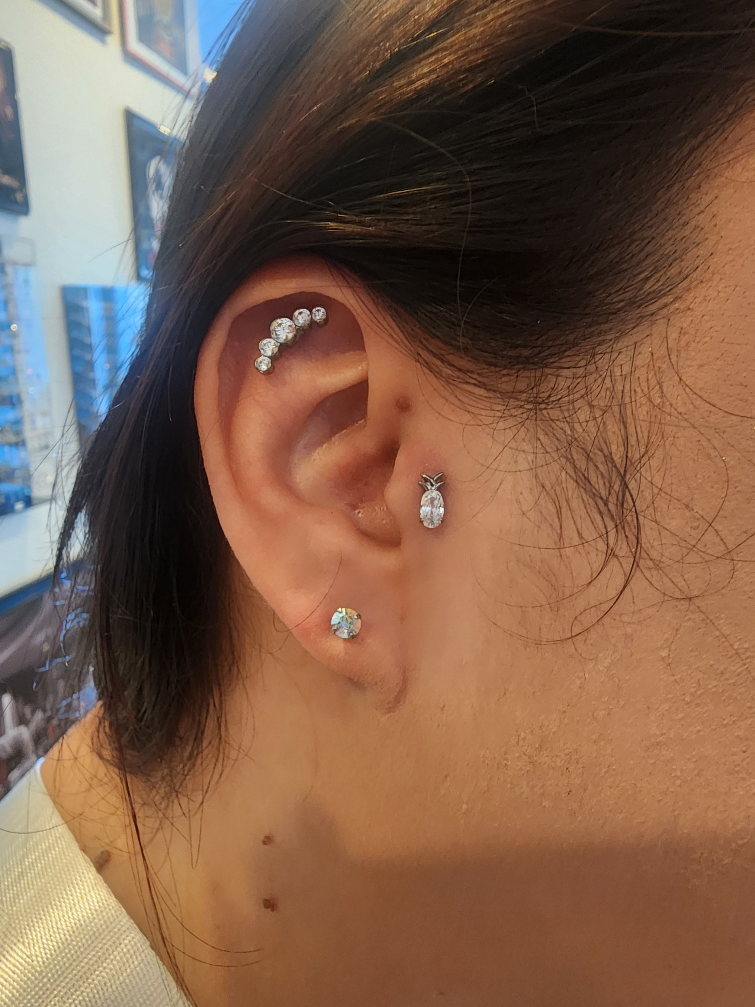 Close-up of a woman's ear wearing multiple earrings, including a curved row of diamonds, a dangling diamond earring, and a small diamond stud, with dark hair and a blurred background.