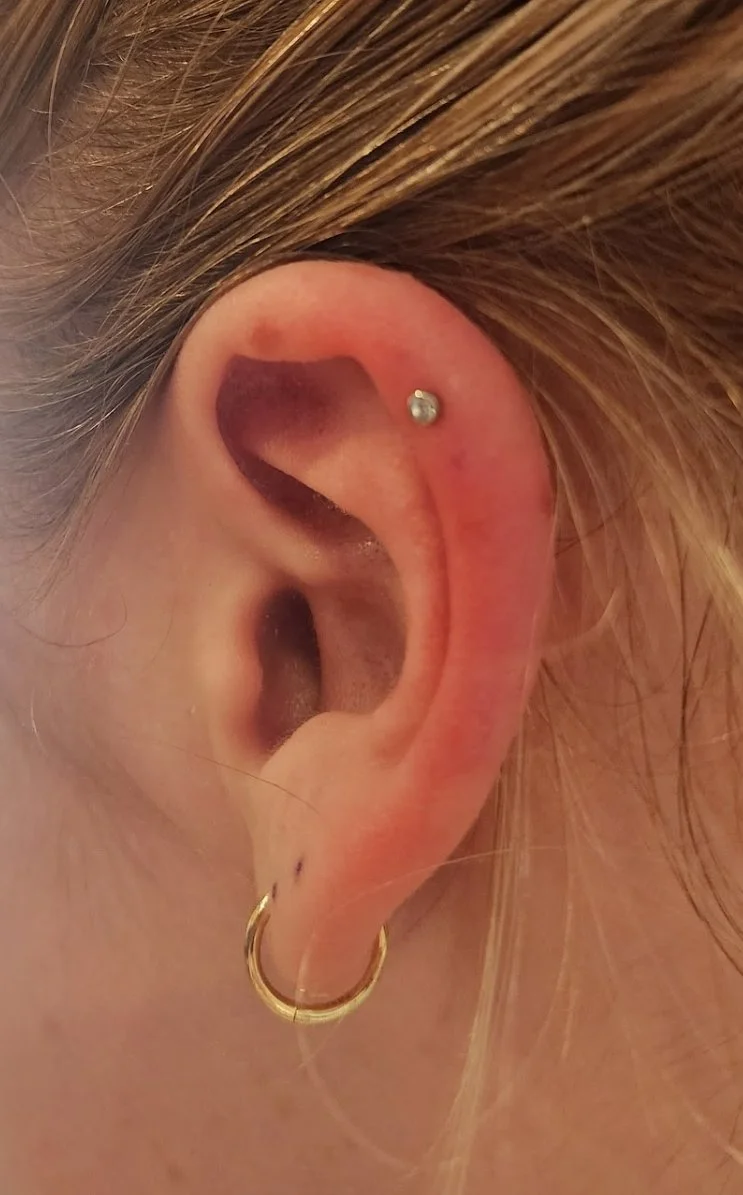 Close-up of a person's ear with multiple piercings, including a silver stud on the upper cartilage and a gold hoop earring in the earlobe, with light brown hair in the background.