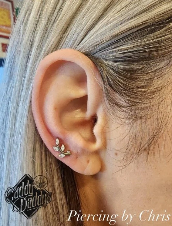 Close-up of a person's ear with a decorative silver and gemstone earring and platinum blonde hair. The image includes a logo that says 'Caddy Dadding' and text that reads 'Piercing by Chris.'