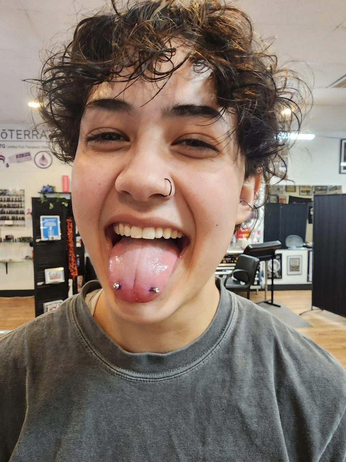 Person with short, curly hair sticking out tongue, showing tongue piercings, smiling with one eye slightly squinted, wearing a nose ring and t-shirt, in a retail or cafe setting.