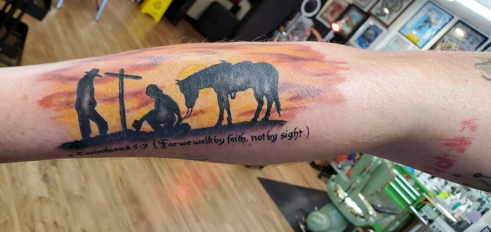 Tattoo of a biblical scene with three silhouette figures, a man by a cross, another kneeling, and an elephant, with a sunset background and a Bible verse referencing 2 Corinthians 5:7.