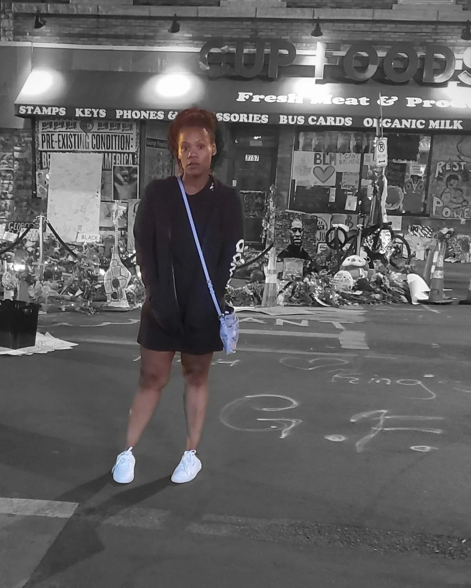 A black woman standing in front of a store with signs, posters, and memorial items, with chalk writing on the ground in her vicinity. Lakeisha Lee MN George Floyd Square