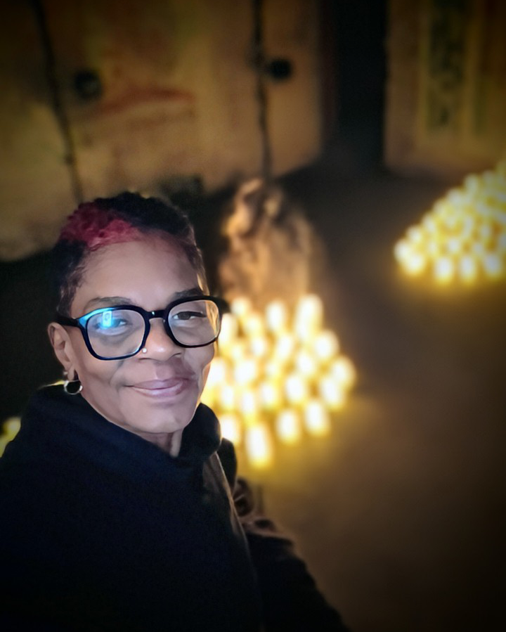 A Black Women Person taking a selfie in a dimly lit space with glowing candles arranged on the floor behind them. 