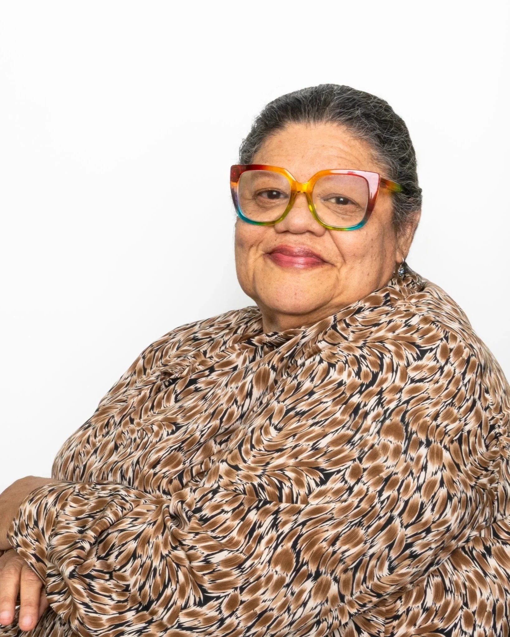 A woman with dark hair pulled back, wearing large rainbow-colored glasses, a leopard print top, and earrings, sitting against a plain white background.