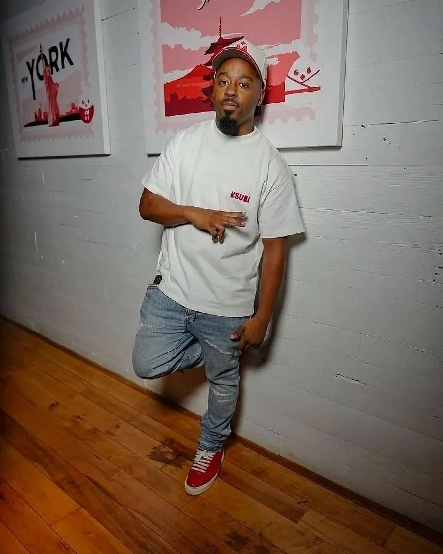 A handsome man standing against a white wooden wall, wearing a white T-shirt with red text, blue jeans, red sneakers, and a gray and red cap, making a hand gesture. 