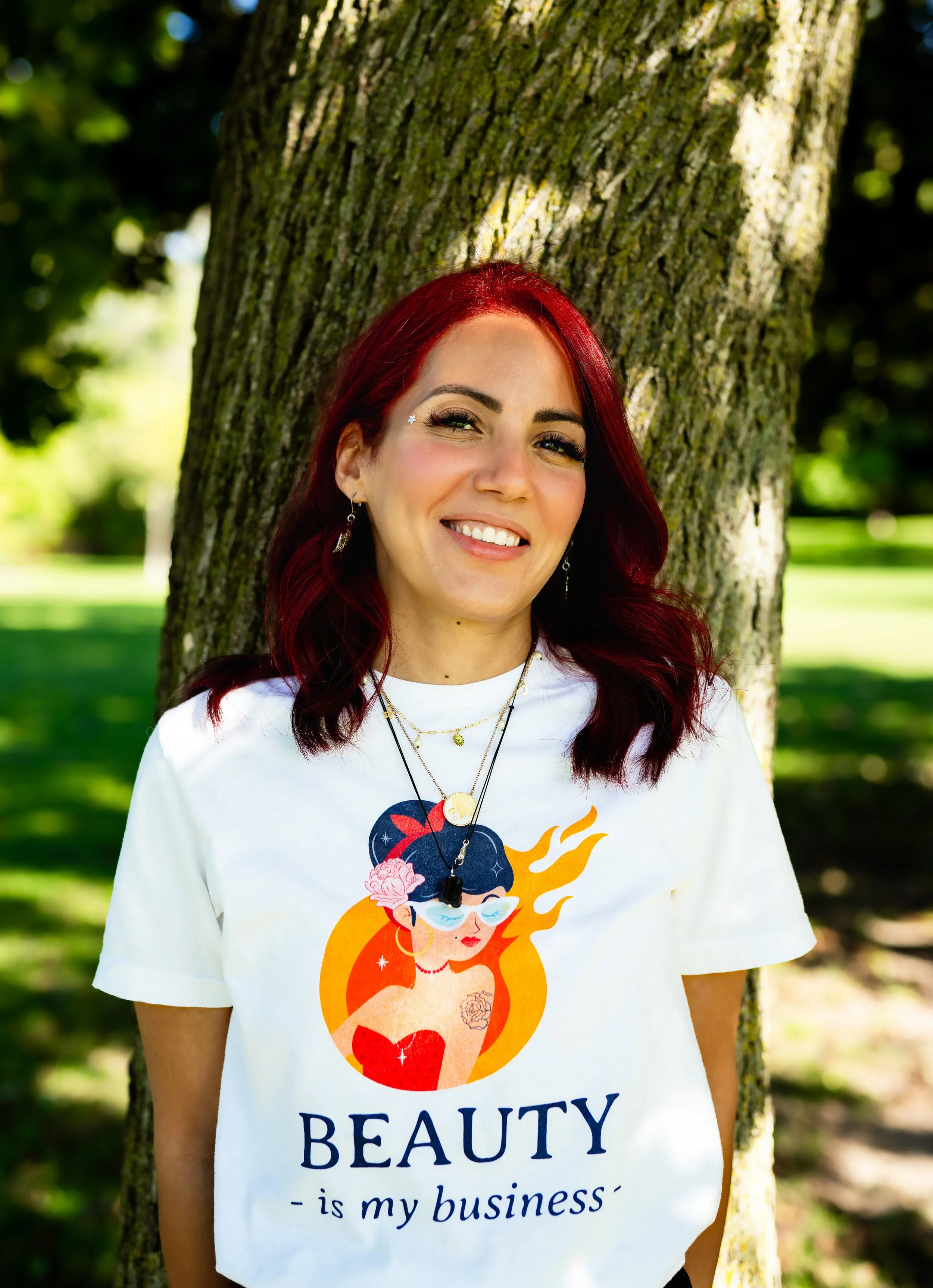 owner and holistic aesthetician outdoors in front of a tree, wearing a white t-shirt with a graphic of a woman with glasses and tattoos, and the text 'Beauty is my business'.