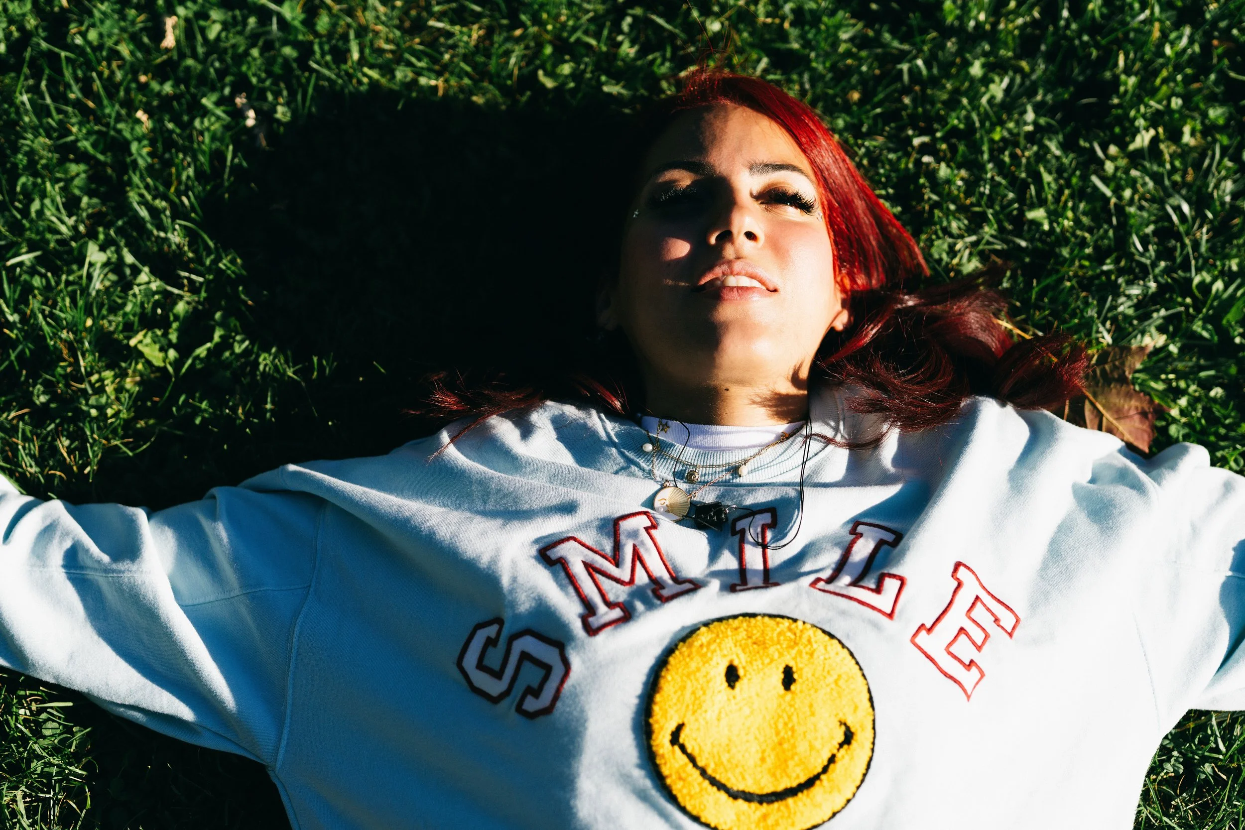 A young woman with red hair lying on grass, wearing a white sweatshirt with a smiley face and the word 'SMILE' on it, and jewelry around her neck.