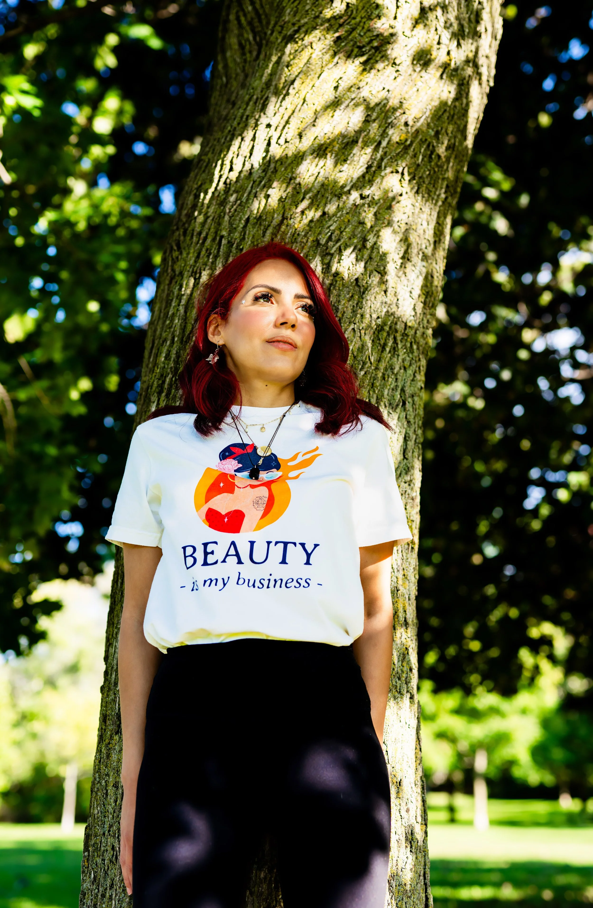A woman with red hair leaning against a tree in a park, wearing a white T-shirt that says 'BEAUTY is my business' with a colorful graphic, and black pants.