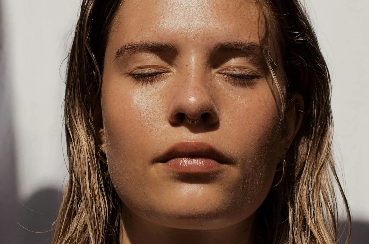 A close-up of a woman's face with her eyes closed, showing wet, textured skin and strands of damp hair, against a blurred background.
