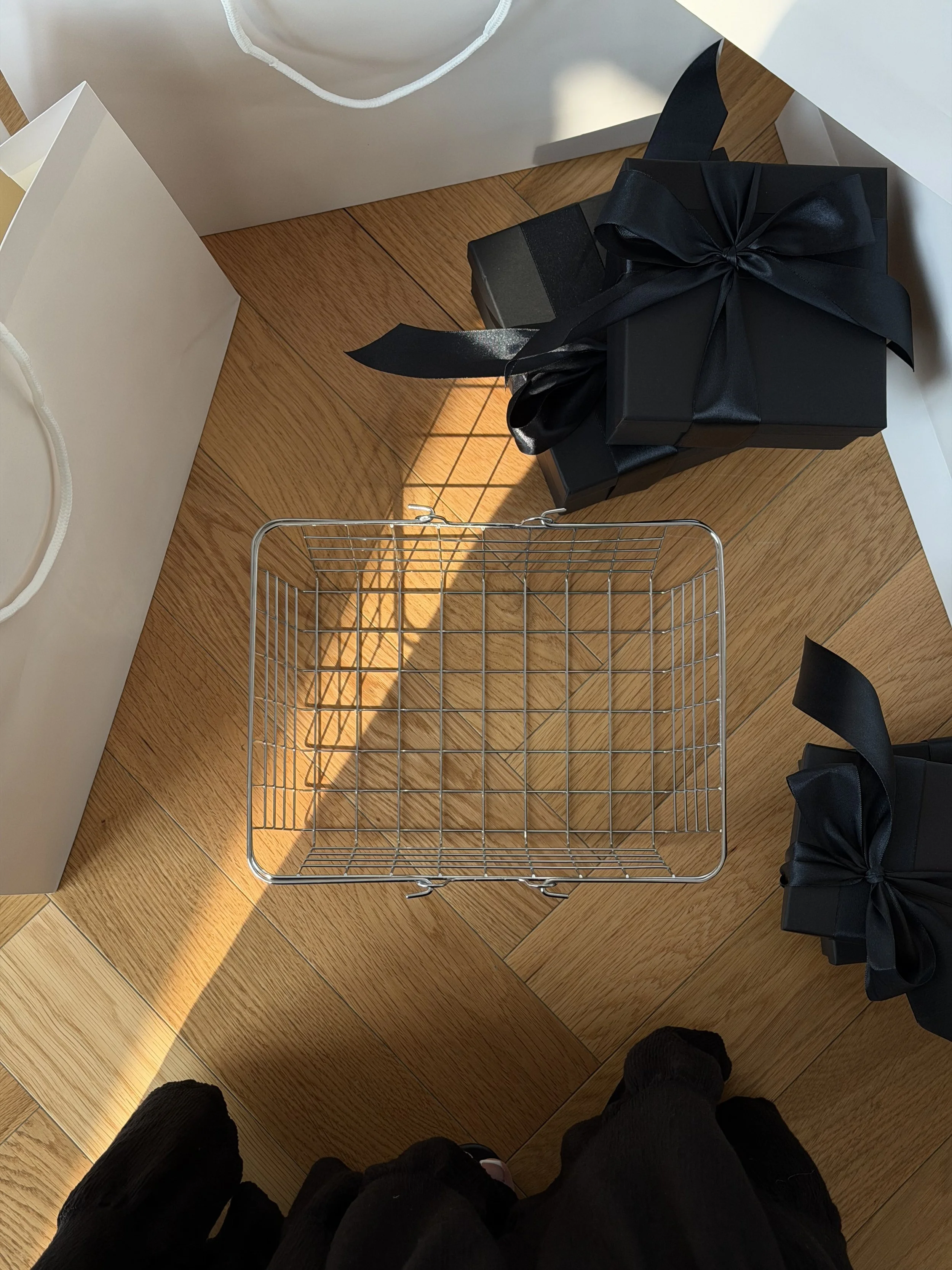 Flat lay of black gift boxes and an empty wire basket on a sunlit wooden floor