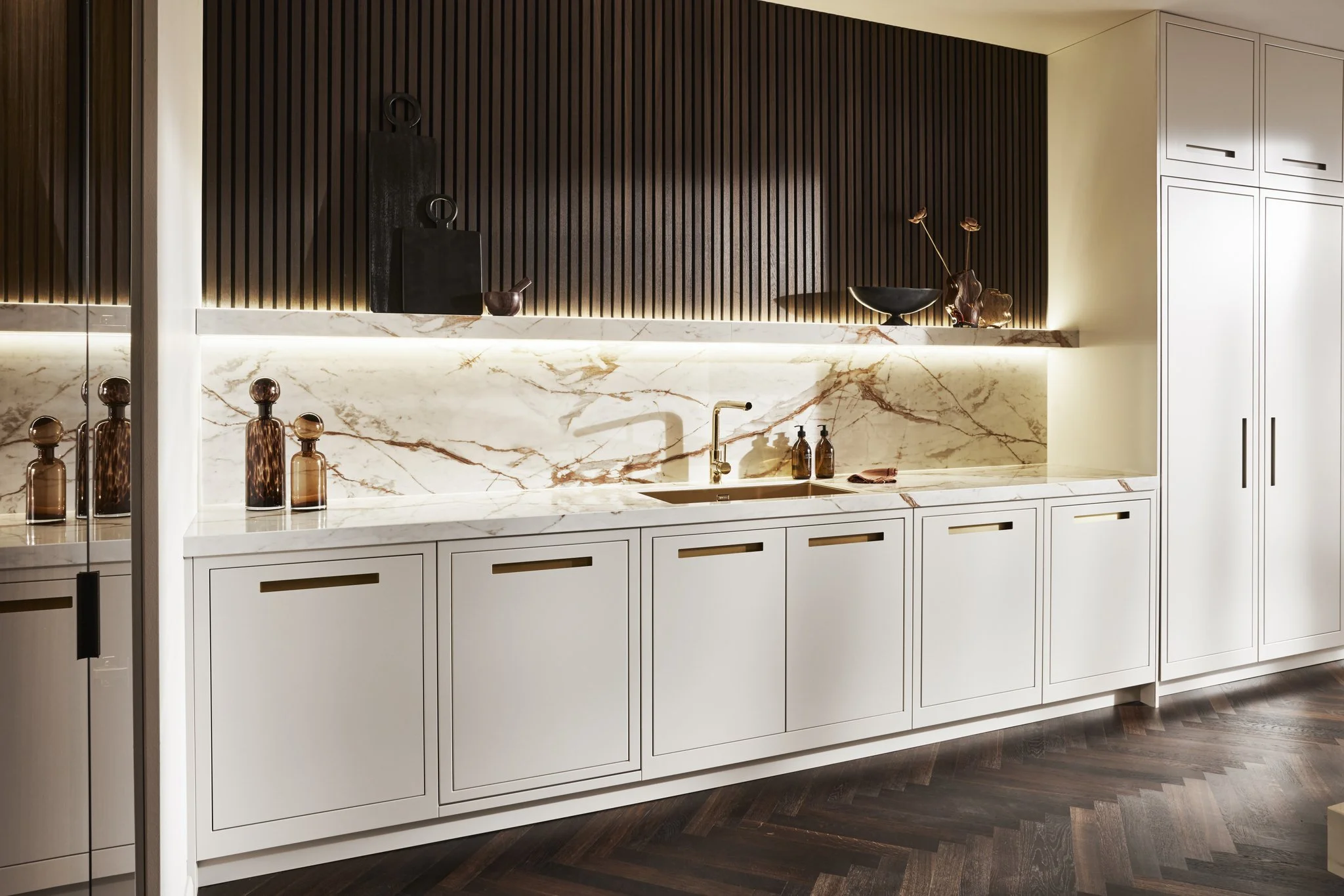 Modern kitchen interior with white cabinets, marble backsplash, and countertop. Features gold faucet, brown decorative bottles, dark wood paneling, and ambient lighting. Brown herringbone wood floor.