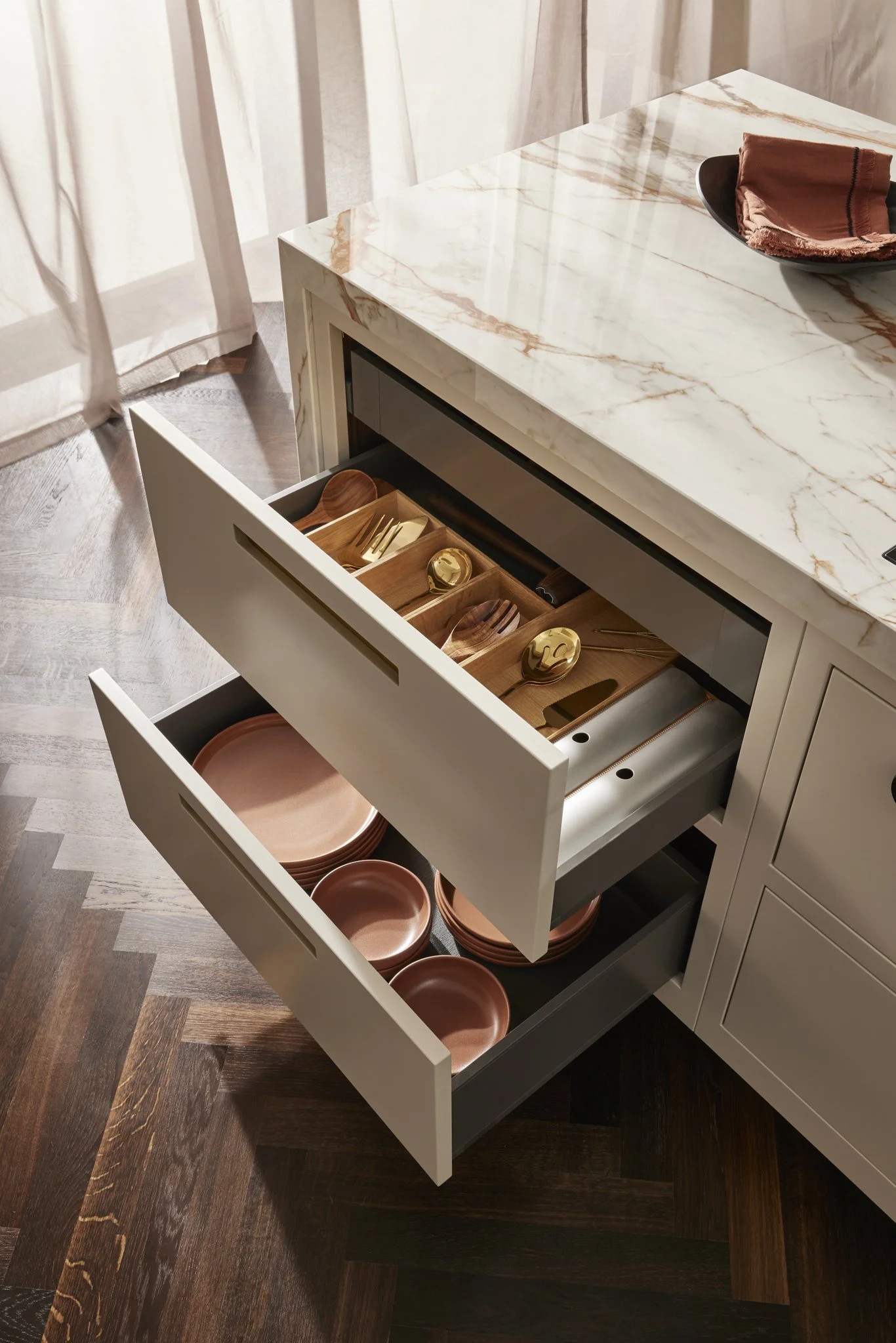 Kitchen island with marble countertop, open drawers showing wooden organizers, gold utensils, and brown ceramic dishes, wood herringbone floor.