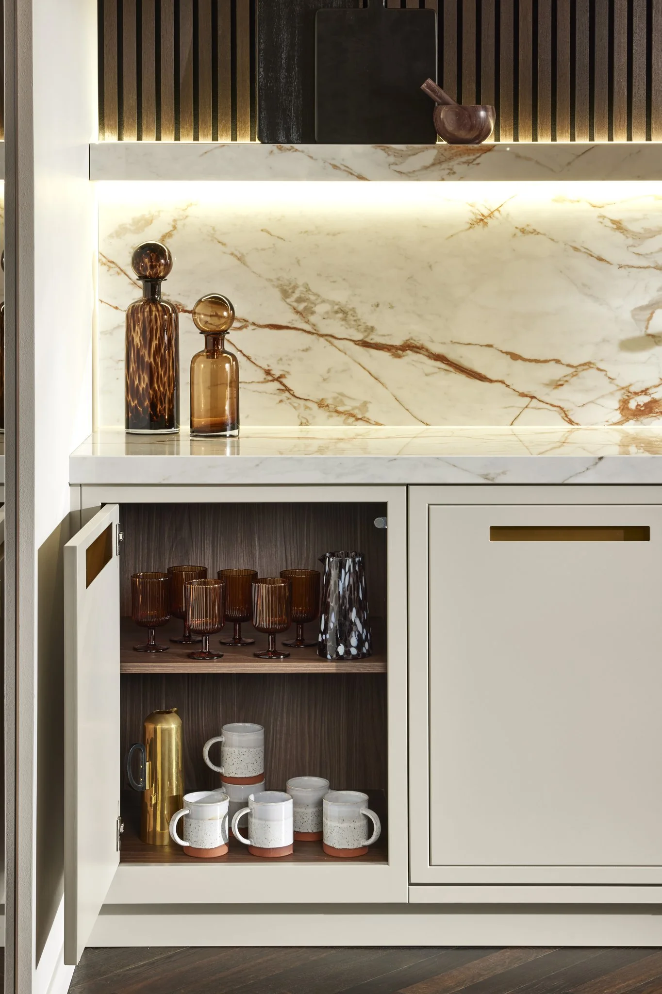 Modern kitchen cabinet with marble countertop, open cupboard displaying glassware and mugs, decorative bottles and wood accents on top.
