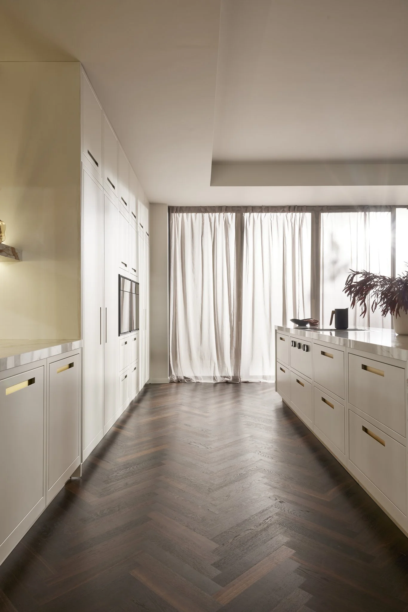 Modern kitchen interior with white cabinetry, dark herringbone wood floor, large windows with sheer curtains, and minimalist decor.
