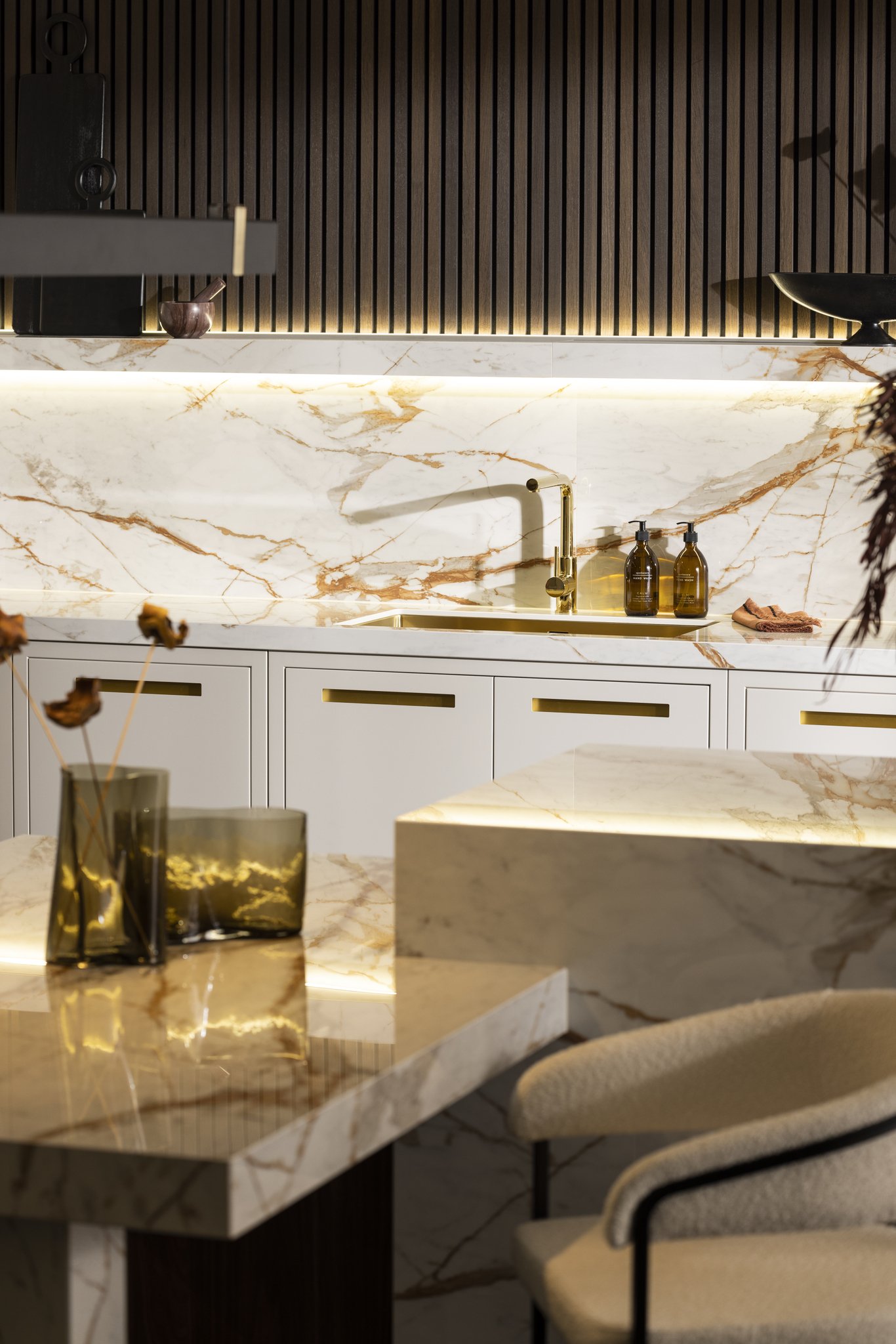 Modern kitchen with marble countertops and backsplash, gold faucet, and glass vases on counter.