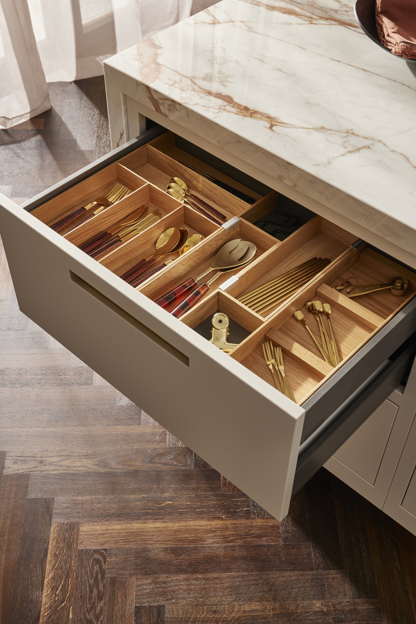 Open kitchen drawer with wooden organizer containing gold-plated cutlery, utensils, and accessories on a wooden floor.