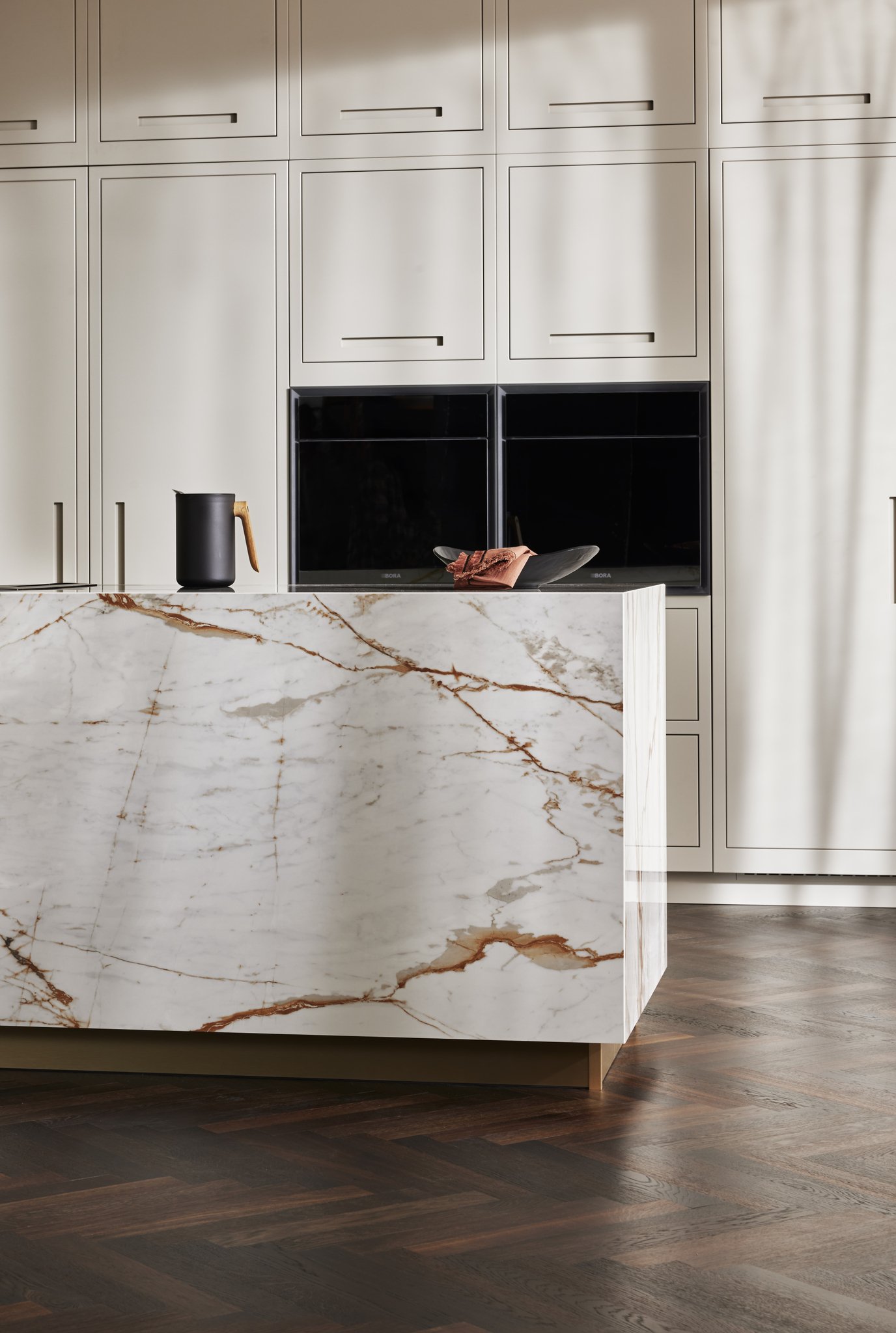 Modern kitchen interior with marble island countertop, black pitcher, wooden cabinets, and dark herringbone floor.