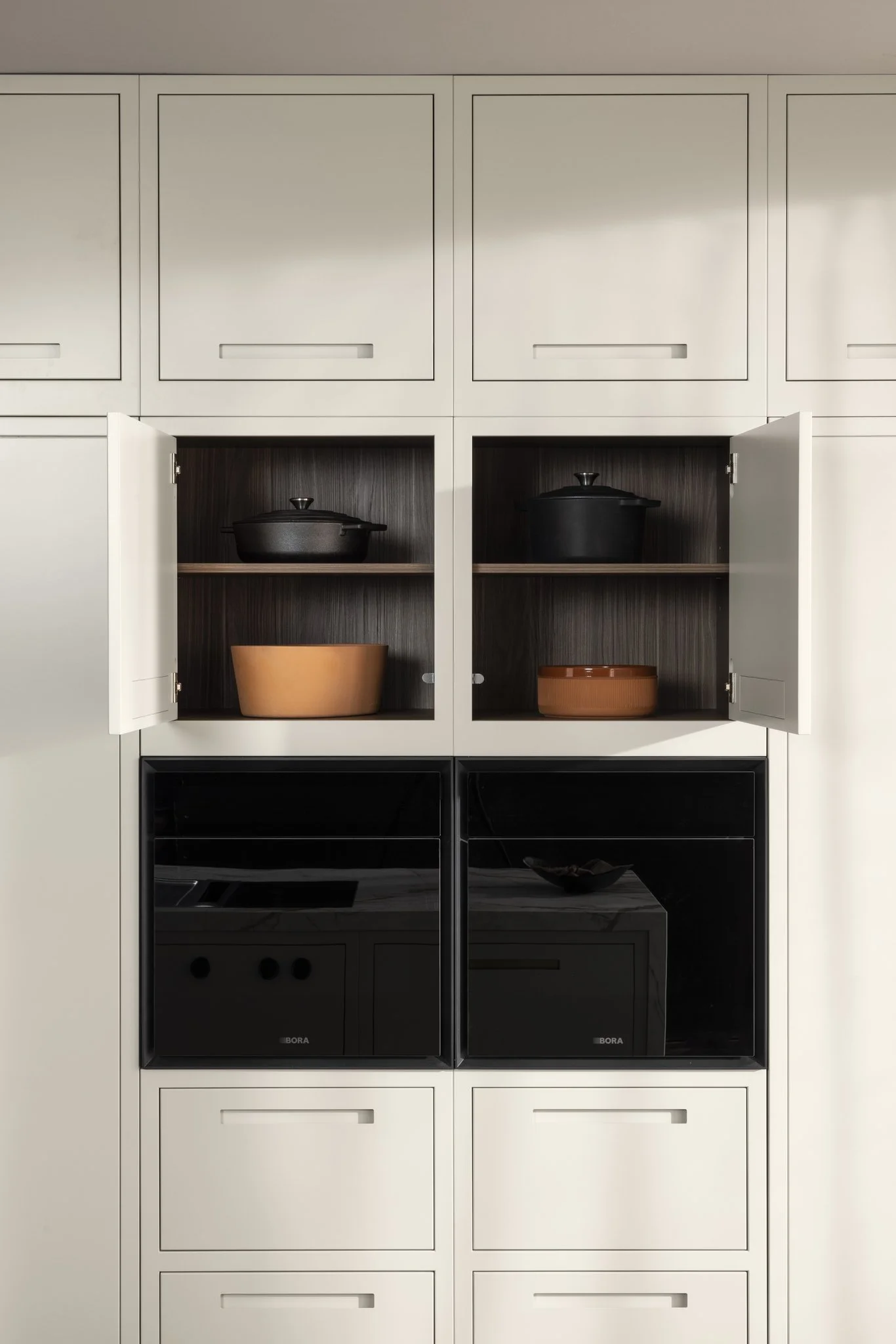 Modern kitchen cabinet with open doors, displaying black cast iron pots and terracotta bowls on wooden shelves.
