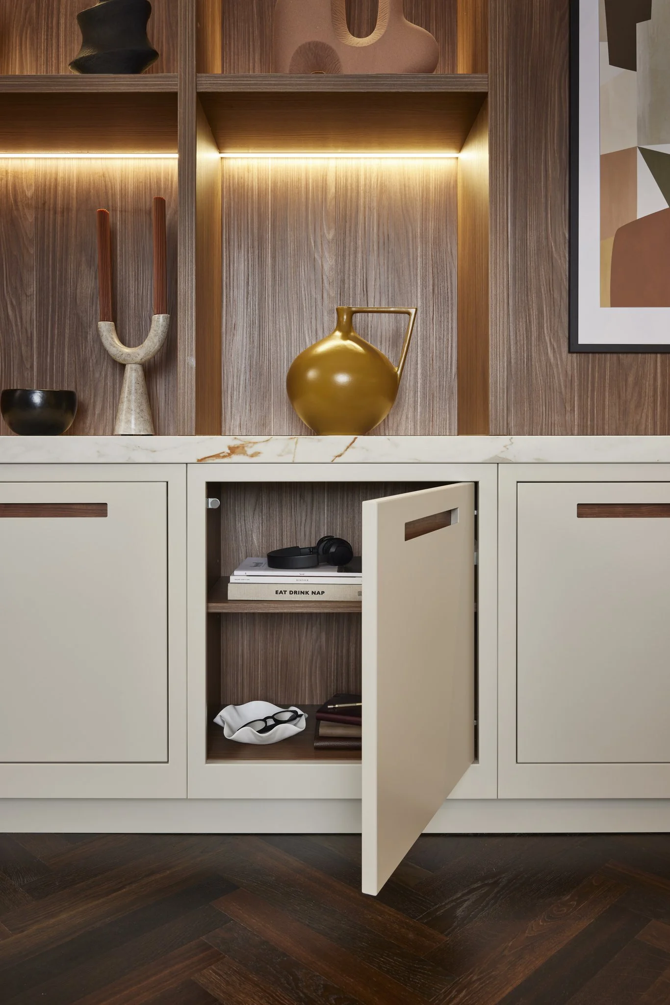 Modern kitchen cabinet with open door, containing books and headphones, decorated with a yellow jug on top, surrounded by abstract art, wooden shelves, and decorative items.