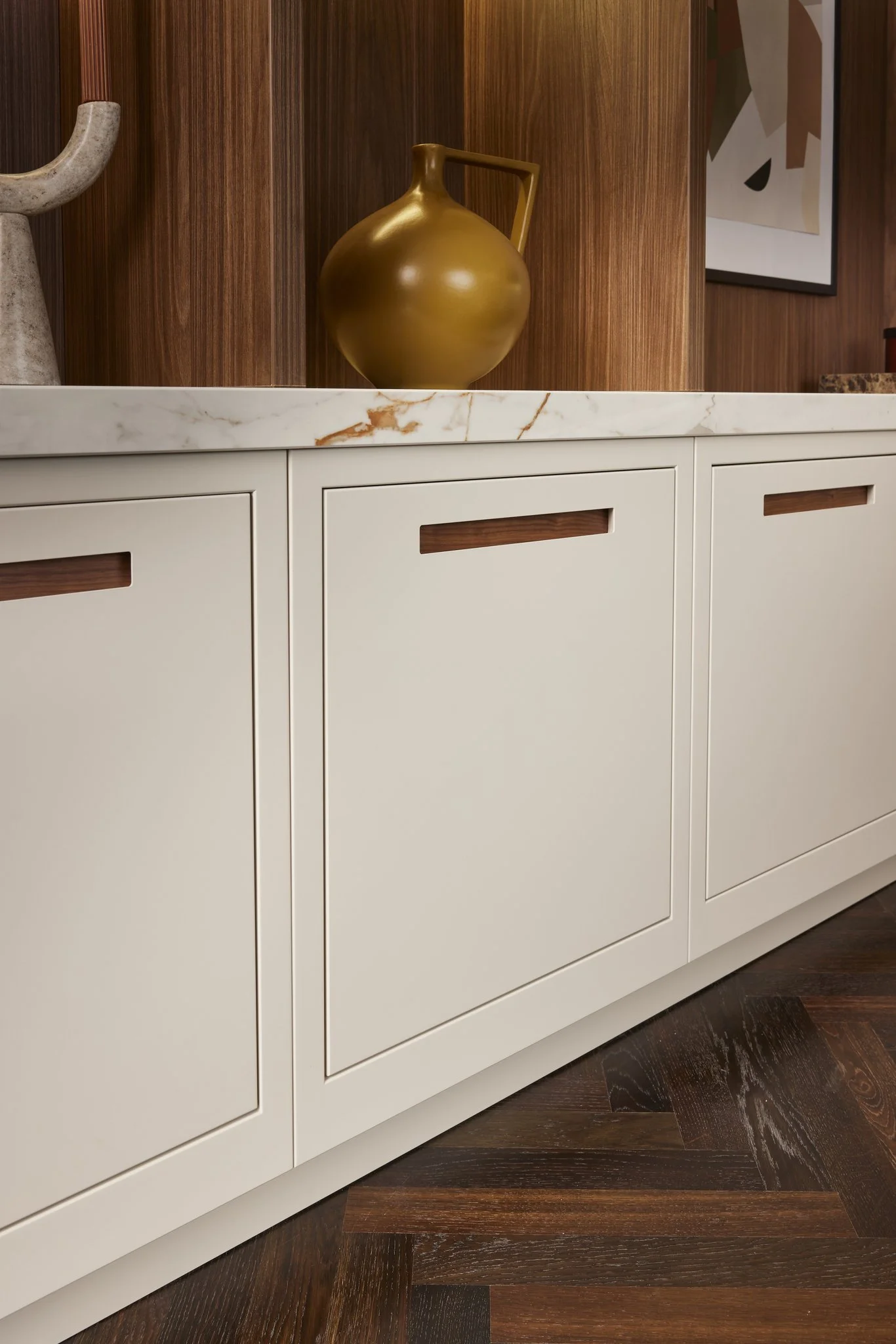 Modern kitchen cabinets with light beige finish, marble countertop, wooden handles, decorative jug, abstract wall art, and parquet wood flooring.