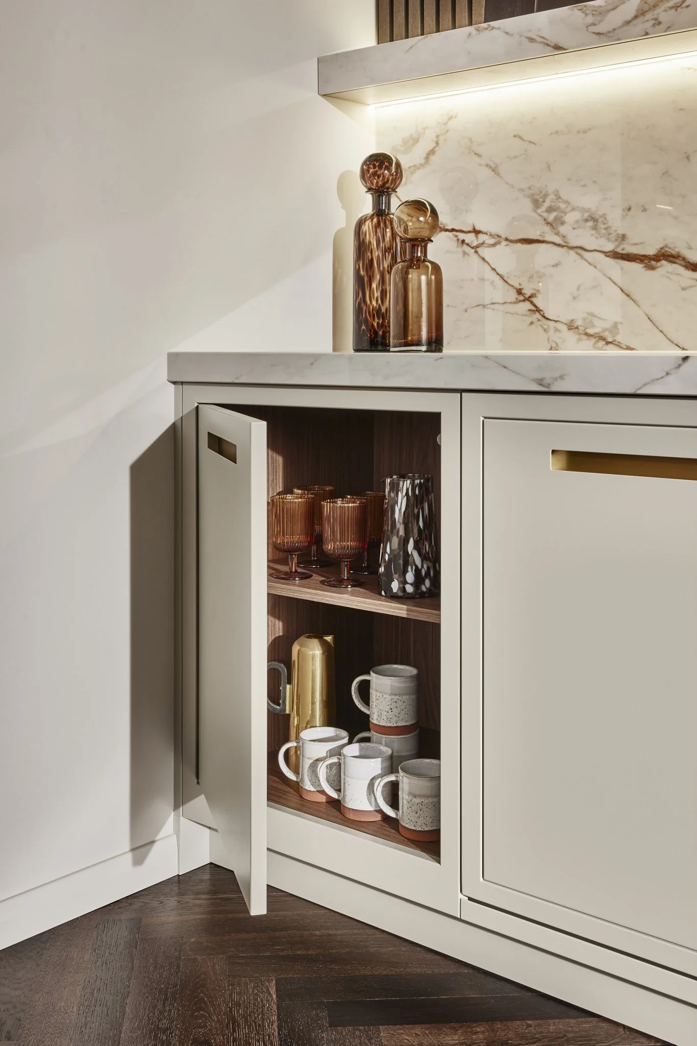 Modern kitchen corner with marble backsplash, featuring two decorative glass bottles on the countertop. A partially open cabinet below reveals shelves with glassware, mugs, and a gold pitcher.