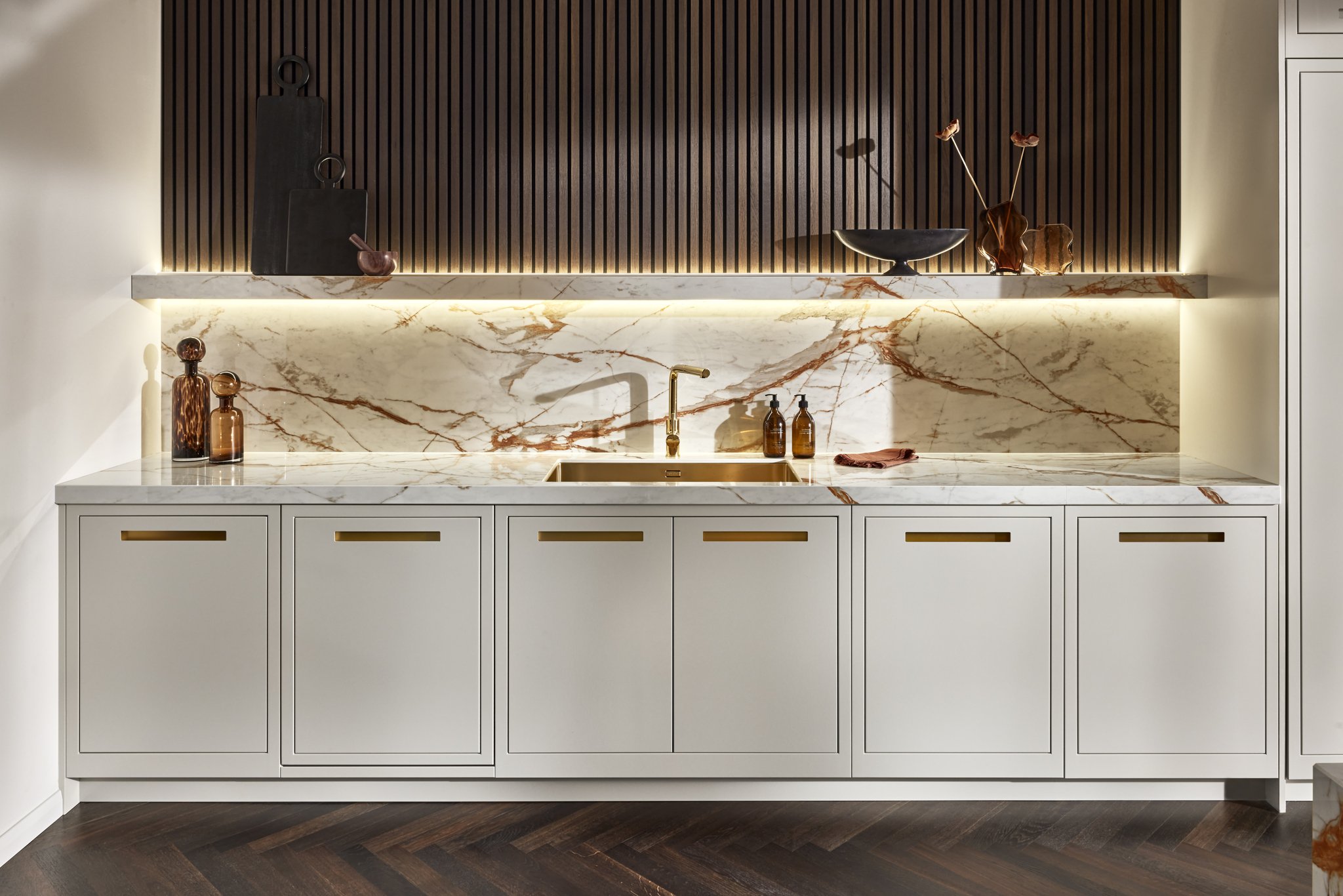 Modern kitchen with marble countertops and backsplash, gold faucet, white cabinets, black wood paneling, and decorative items.