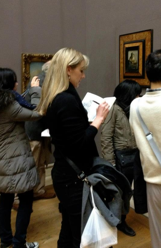 People observing artwork in a museum with art frames on the wall and a woman reading a pamphlet.