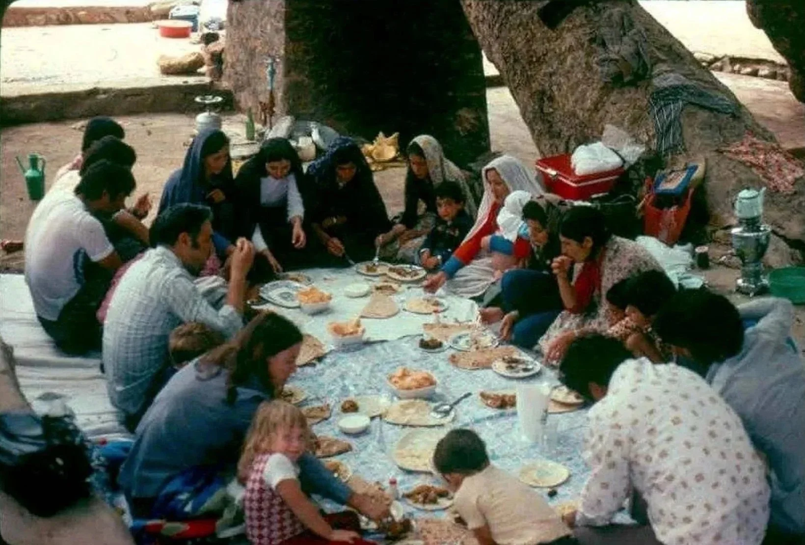 People gathered on the ground under a tree, enjoying a meal together outdoors, with various dishes and plates on a tablecloth.
