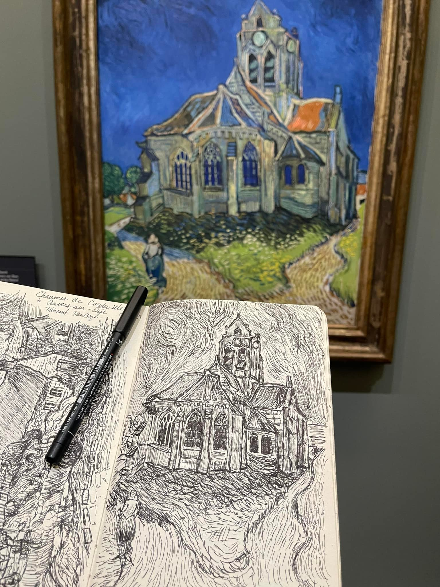 A painting of a church with a blue sky background, framed on a wall. In the foreground, there's a sketchbook open with a black ink drawing of the same church, featuring detailed lines and shading, accompanied by a black pen resting on it.