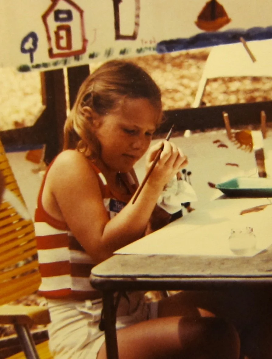 A young girl with blonde hair is sitting at a table, looking at a piece of paper with a pencil in her hand. She appears focused or concentrated on her work.